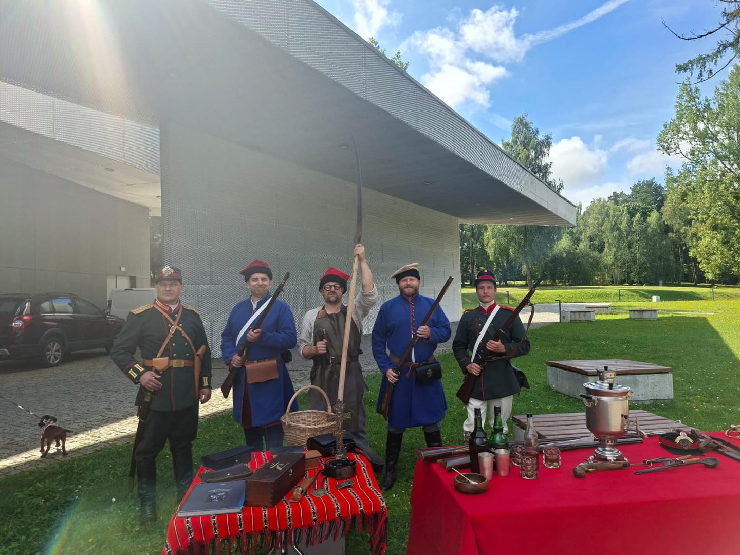 Na zdjęciu widać pięciu mężczyzn w historycznych strojach wojskowych, stojących przed nowoczesnym budynkiem Muzeum Bitwy pod Grunwaldem. Trzymają broń. Na pierwszym planie znajduję się stół przykryty czerwonym materiałem, na którym ustawiono rekwizyty związane z epoką Powstania Styczniowego: księgi, skrzynki, butelki, kieliszki, naczynia i samowar. W tle widać drzewa i trawnik oraz fragment zaparkowanego samochodu i osobę z psem.